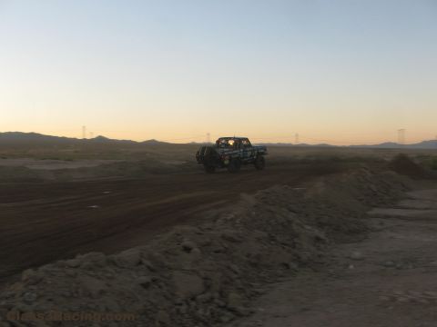 1990 Ford Bronco - off-road race truck travels along a desert dirt road at dusk with transmission towers and a fading sunset behind.