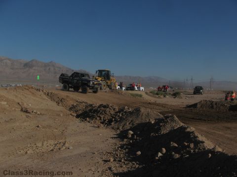 1990 Ford Bronco - front loader pushes an off-road race truck up a dirt berm while spectators and vehicles gather along a desert course under blue sky.