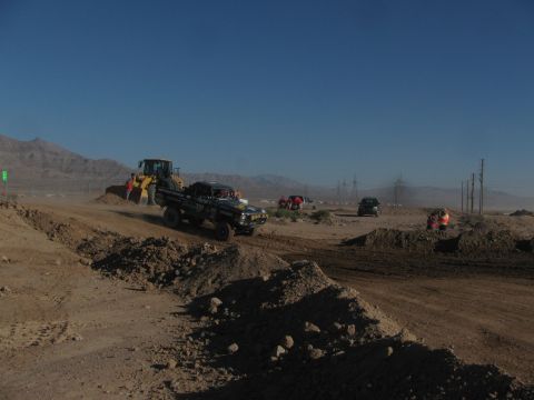 1990 Ford Bronco - wheel loader assists a dark off-road race truck over a dirt berm as crews and vehicles line the desert course under blue sky.