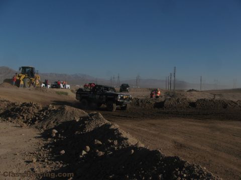 1990 Ford Bronco - dark off-road race truck drives a desert dirt course past a wheel loader and crews in safety vests under a clear blue sky.