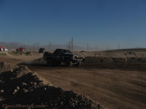 1990 Ford Bronco - dark off-road race truck rounds a banked dirt berm on a desert course with mountains and power lines under blue sky.