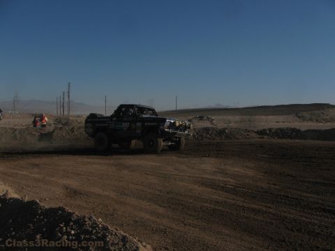 1990 Ford Bronco - number 300 off-road race truck drives a desert dirt course with a marshal and power lines visible under a clear blue sky.