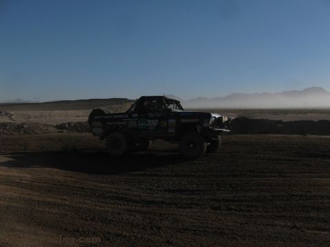 1990 Ford Bronco - number 300 West Coast Broncos race truck crosses a dusty desert dirt course with a spare tire on the bed and distant mountains.