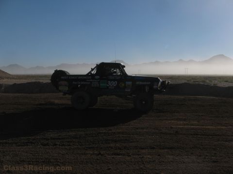 1990 Ford Bronco - number 300 off-road race truck is silhouetted amid dust haze on a desert dirt course with mountains in the background.