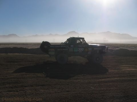 1990 Ford Bronco - West Coast Broncos number 300 race truck is backlit against dust haze on a desert dirt course with mountains behind.
