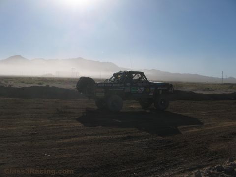 1990 Ford Bronco - off-road race truck numbered 300 kicks up dust as it drives across a hazy desert course backed by distant mountains.