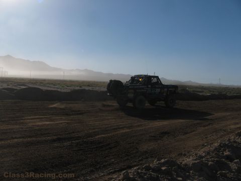 1990 Ford Bronco - backlit off-road race truck moves along a dusty desert course with mountains and morning haze in the background.
