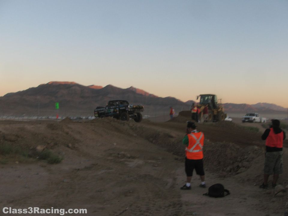 Race truck on a dirt berm at dusk with safety crew and a wheel loader nearby