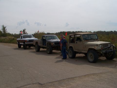 1989 Ford Bronco - Three modified trucks and SUVs are lined up on a dirt road, possibly at an off-road event.