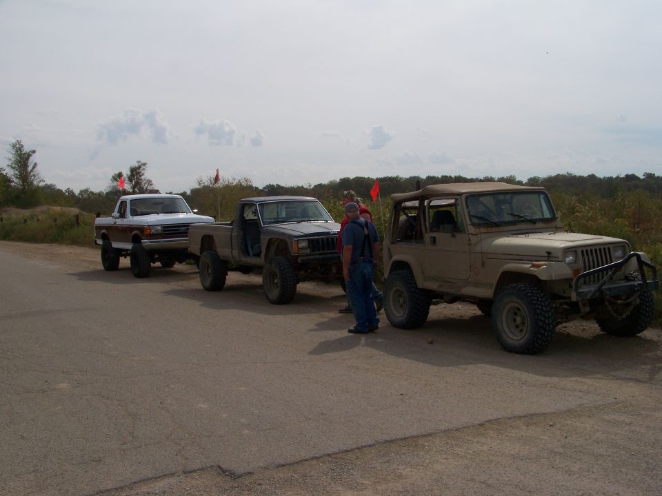 Tan Jeep Wrangler with off-road modifications, parked behind a pickup truck