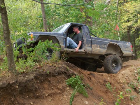 1989 Ford Bronco - A man drives a Jeep Comanche pickup truck up a steep, wooded trail.
