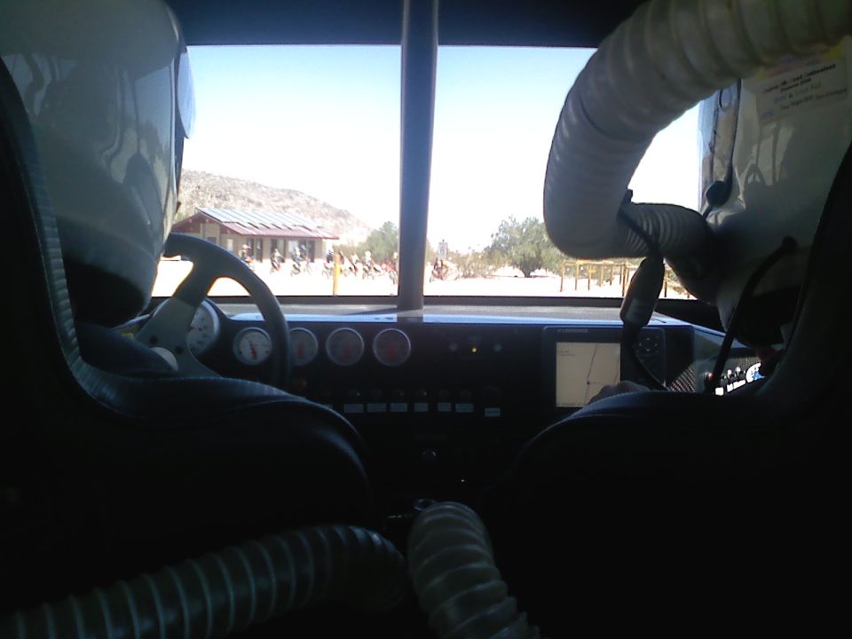 Interior view from driver's seat of a modified vehicle, looking out at a desert landscape with a building