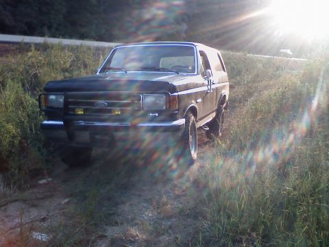 1990 Ford Bronco - A Ford Bronco sits off-road as the sun sets, casting lens flare across the image.