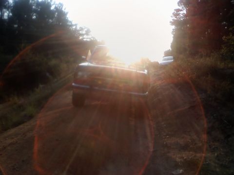 1990 Ford Bronco - A dark SUV travels down a dusty road, with another vehicle visible in the distance and the sun creating lens flare.