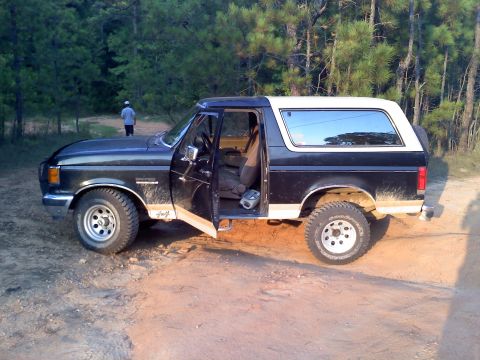 1990 Ford Bronco - This 1990 Ford Bronco is parked on a dirt path surrounded by trees, with the driver's door ajar.