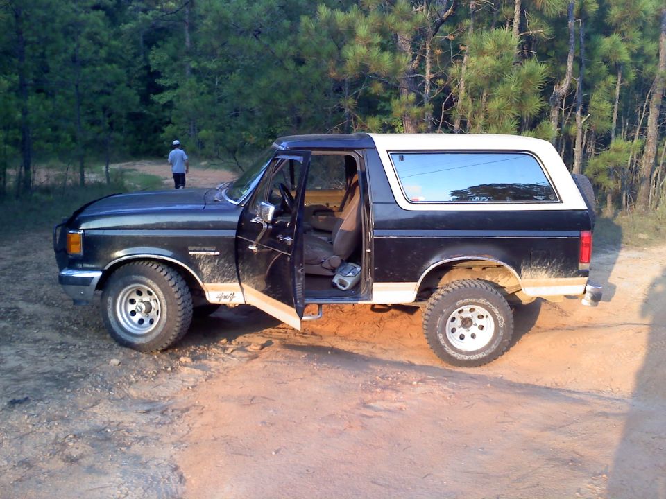 1990 Ford Bronco parked on dirt road with trees in background, driver door open