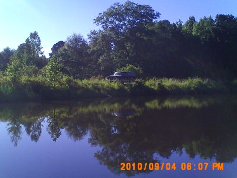 1990 Ford Bronco - A dark-colored SUV is parked on the edge of a grassy bank next to a calm body of water, with trees in the background.