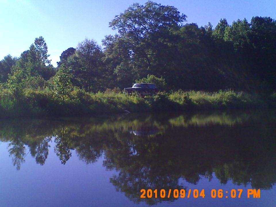 Dark SUV parked on grassy bank beside a lake, reflected in water