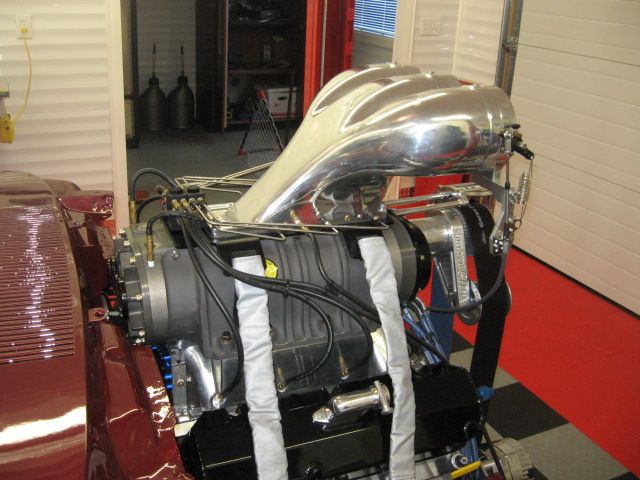 Roots supercharger with polished scoop and blower straps on V8 in dark red car, hood off