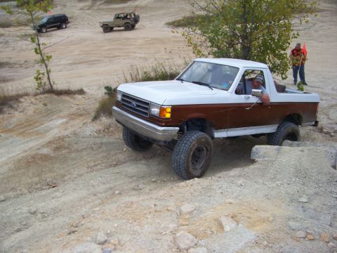 1989 Ford Bronco - A two-tone Ford Bronco navigates a challenging off-road course with other vehicles in the background.