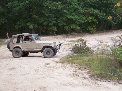 1989 Ford Bronco - A tan Jeep Wrangler is driven on a dirt path through a wooded area.