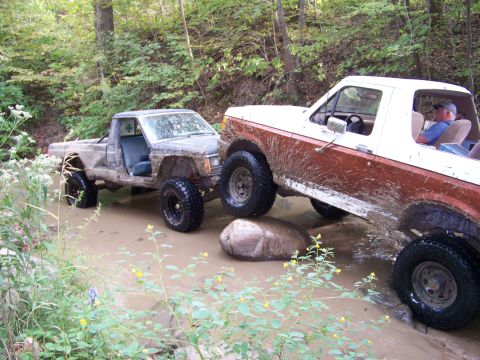 1989 Ford Bronco - Two off-road vehicles navigate a muddy stream bed surrounded by trees.