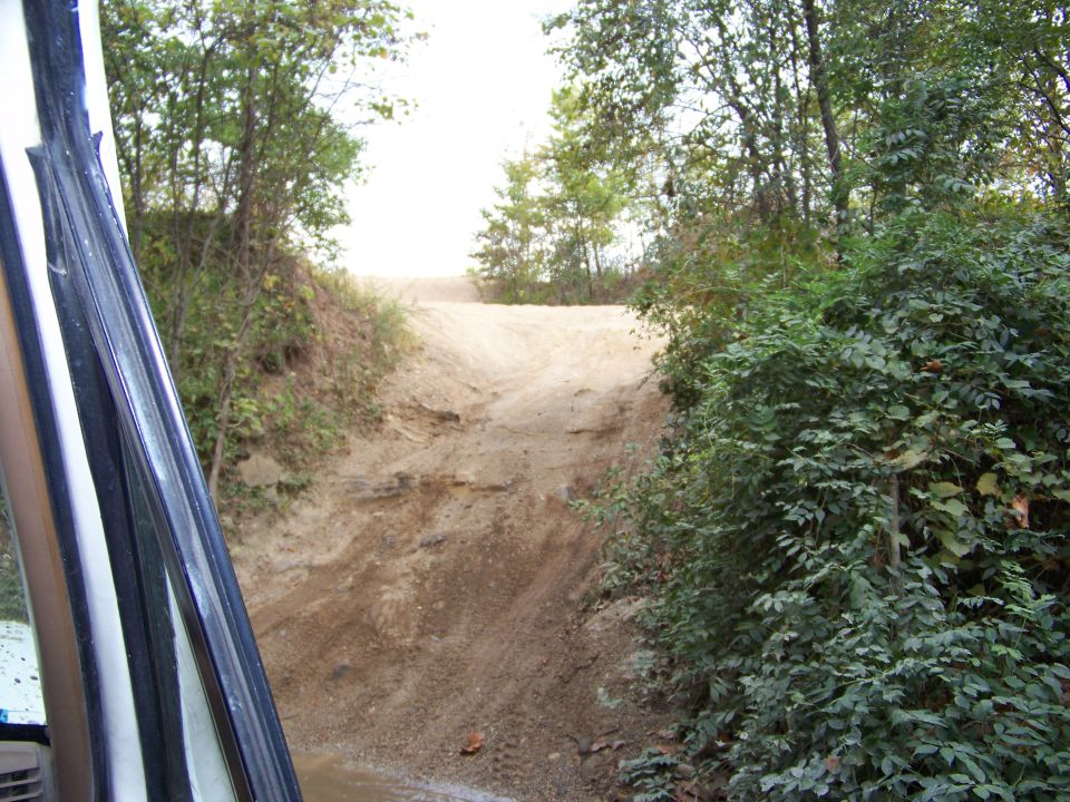 View from inside a vehicle looking out at a steep dirt trail with trees on either side. Water is visible on the ground