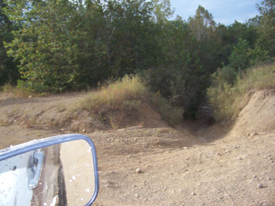Side mirror of a dirty vehicle reflects a dirt road with trees in the background