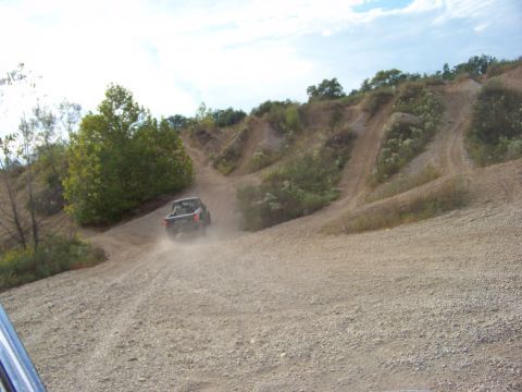 1989 Ford Bronco - A dark lifted 4x4 pickup climbs a dusty gravel hill at an off-road park, kicking up a dust trail as it heads toward eroded trails cut into…