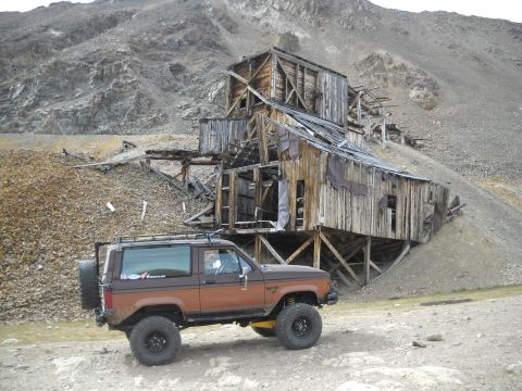 1986 Ford Bronco II - A brown 1986 Ford Bronco II is parked in a , mountainous landscape with an old, dilapidated wooden mine structure in the background.