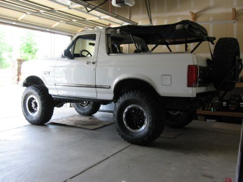 1995 Ford Bronco - White Ford Bronco full-size SUV with removed soft top, viewed from the rear driver's side, parked in a garage. It has a suspension lift, la…