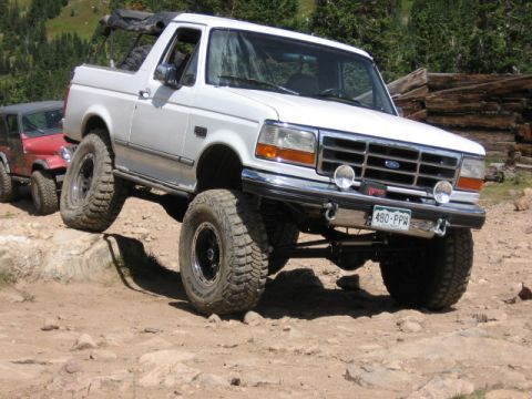 1995 Ford Bronco - A white 1990s Ford Bronco full-size SUV with a lifted suspension and large off-road mud tires, shown from a 3/4 front angle climbing over r…
