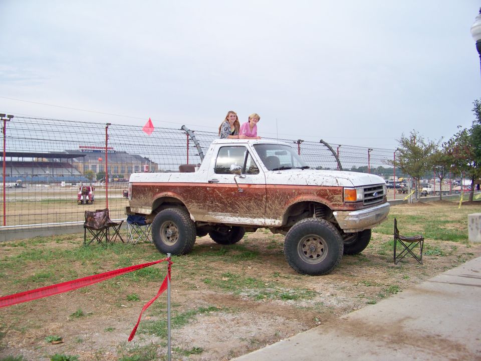 Muddy lifted Ford Bronco with two girls on the roof, parked near a racetrack
