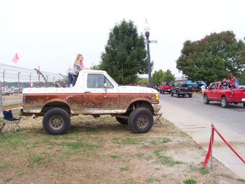 1989 Ford Bronco - A two-tone Ford pickup truck, covered in mud, is parked on the side of a road with other vehicles in the background.