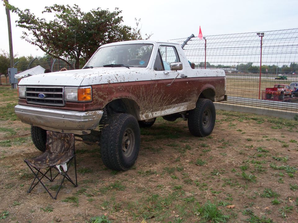 Mud-splattered 1989 Ford Bronco parked on dirt with a camouflage chair in front