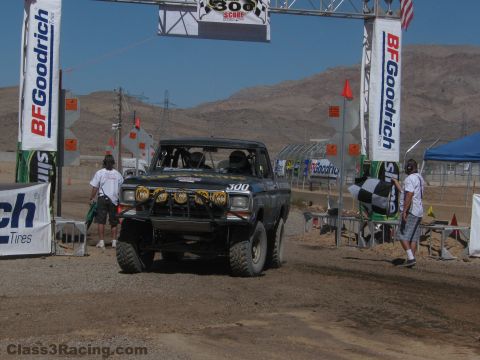 1990 Ford Bronco - black number 300 off-road race truck sits under a BFGoodrich start banner as crew and a checkered flag stand by.