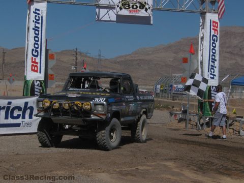 1990 Ford Bronco - number 300 race truck waits under a BFGoodrich banner arch while a man waves a checkered flag on the dirt.