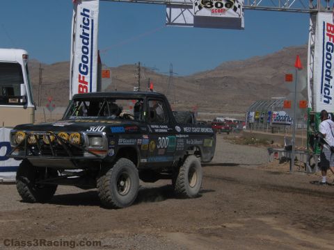 1990 Ford Bronco - black number 300 West Coast Broncos race truck drives through a BFGoodrich banner arch on a desert dirt course.
