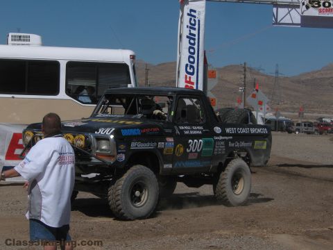 1990 Ford Bronco - number 300 West Coast Broncos race truck rolls through a dirt paddock past a parked bus and an official in a polo.