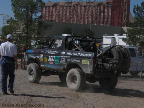 1990 Ford Bronco - number 300 West Coast Broncos race truck drives on gravel as a man walks past with a tall hotel in the background.
