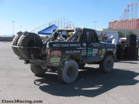 1990 Ford Bronco - Rear view of the number 300 West Coast Broncos race truck with stacked spare tires, parked in a lot near a roller coaster.