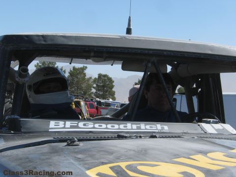 1990 Ford Bronco - Two occupants, one in a white helmet, sit inside the cab of a race truck with a BFGoodrich windshield banner.