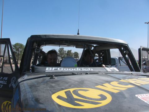 1990 Ford Bronco - Two men sit inside the cab of a race truck whose hood carries a large yellow KC logo and a BFGoodrich banner.