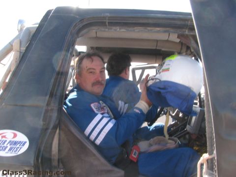 1990 Ford Bronco - mustached man in a blue racing suit sits in a truck cab beside a teammate, holding a white racing helmet.
