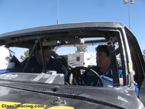 1990 Ford Bronco - Two men in blue racing suits talk inside the open cab of a race truck with a roll cage and steering wheel visible.