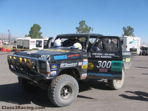 1990 Ford Bronco - number 300 West Coast Broncos race truck sits in a paddock with two helmeted occupants and RVs behind it.