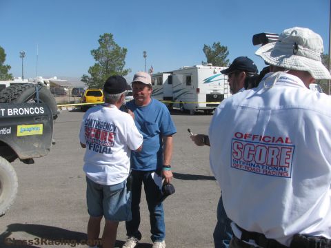1990 Ford Bronco - man in a blue shirt chats with SCORE off-road racing officials in white shirts in a paved paddock near an RV.