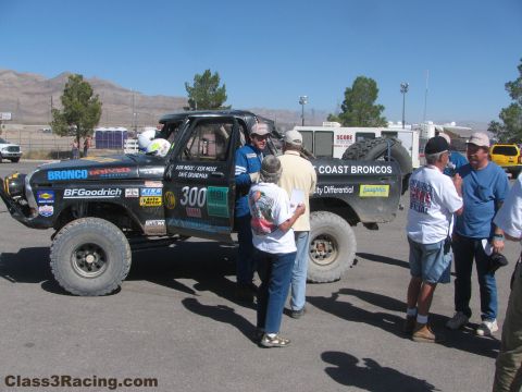 1990 Ford Bronco - small group of people gathers around the number 300 West Coast Broncos race truck in a paved paddock.