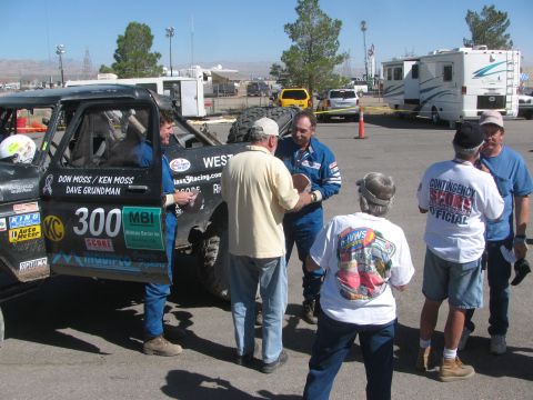 1990 Ford Bronco - Men in blue racing suits and casual clothes converse beside the number 300 West Coast Broncos race truck in a paddock.