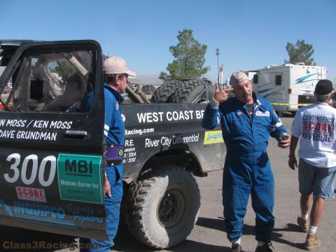 1990 Ford Bronco - Two men in blue racing suits stand beside the number 300 West Coast Broncos race truck, one gesturing, RVs behind.